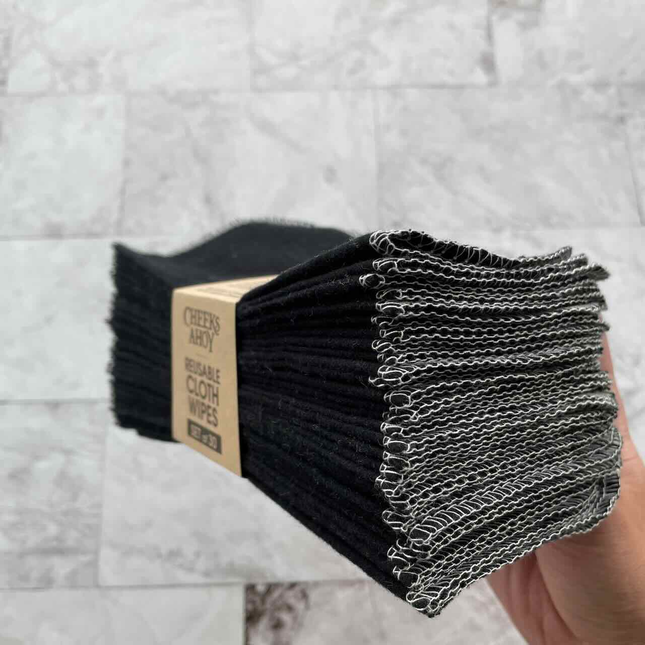 A hand holding dark coloured cheeks ahoy cloth wipes, side view, in its paper sleeve packaging, in a marble patterned background drop.