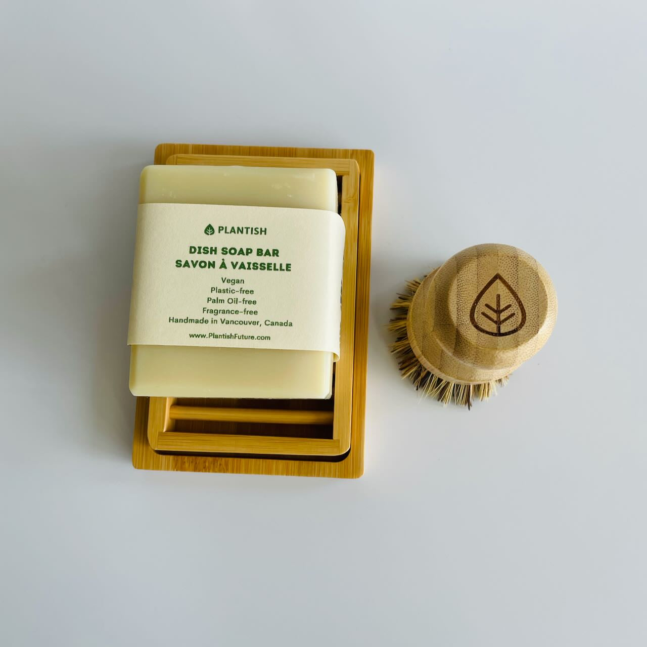 A solid dish soap in its original packaging on a bamboo soap dish, with a sisal bamboo scrubber facing down next to it