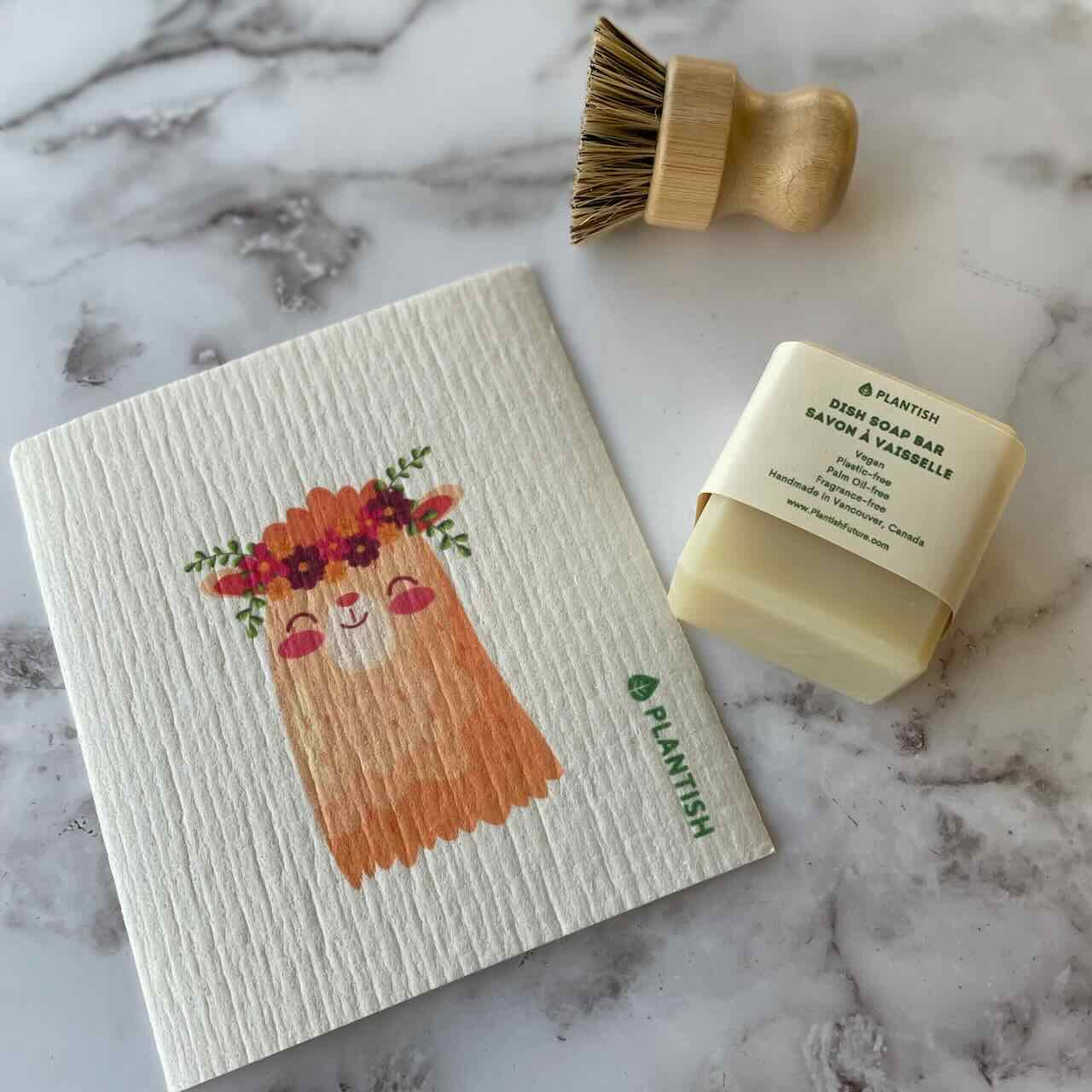 A llama patterned swedish dishcloth lay flat on a marbled countertop. Next to its right, there is a block of solid dish washing soap and a bamboo scrubber.