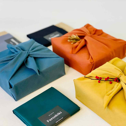 An assortment of gifts wrapped in furoshiki wraps on top of a white table top. Main colours on display are orange, yellow and blue