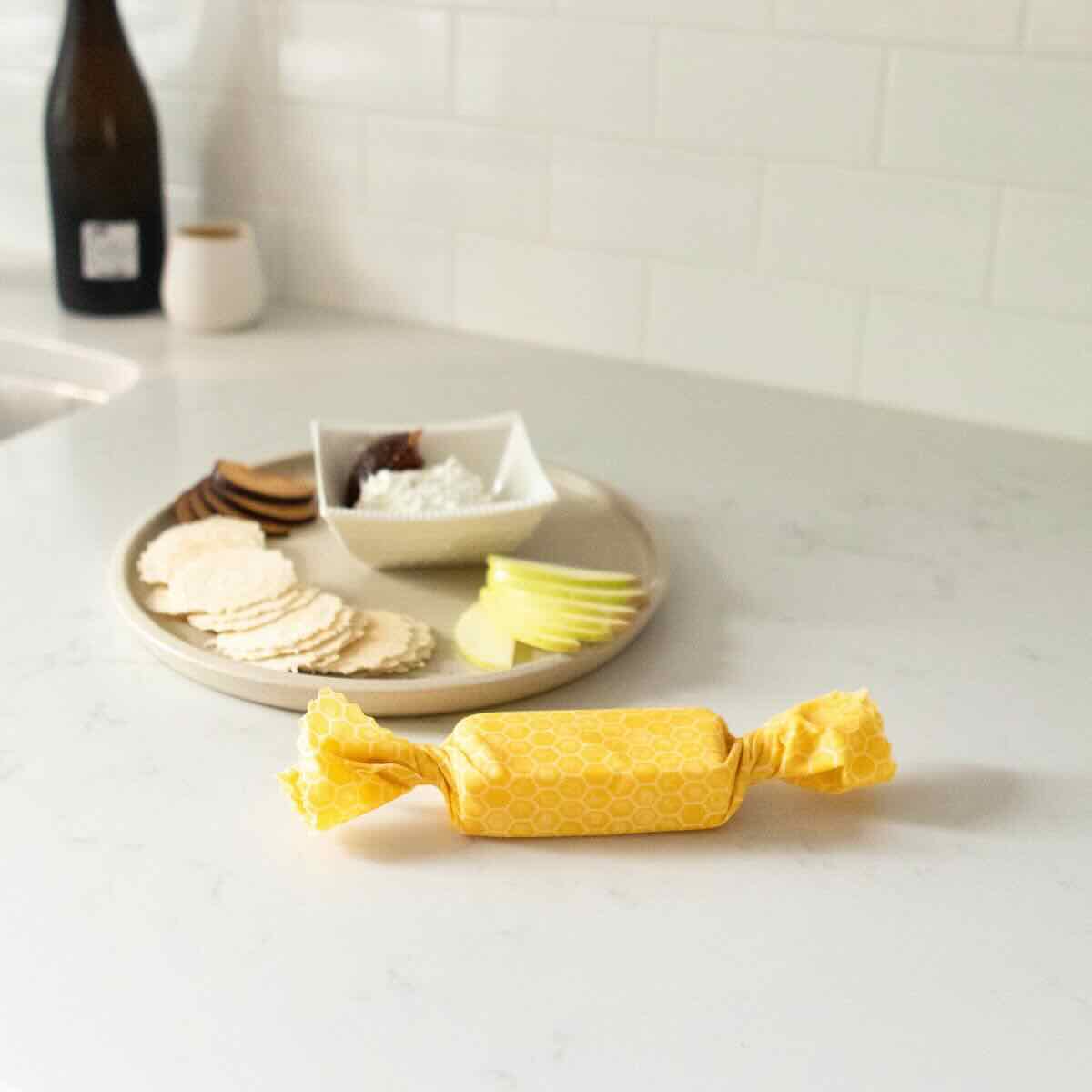 Cheese wrapped in a yellow Goldilocks honeycomb themed beeswax wrap on a concrete kitchen counter top, with cheese and crackers on a plate behind it.