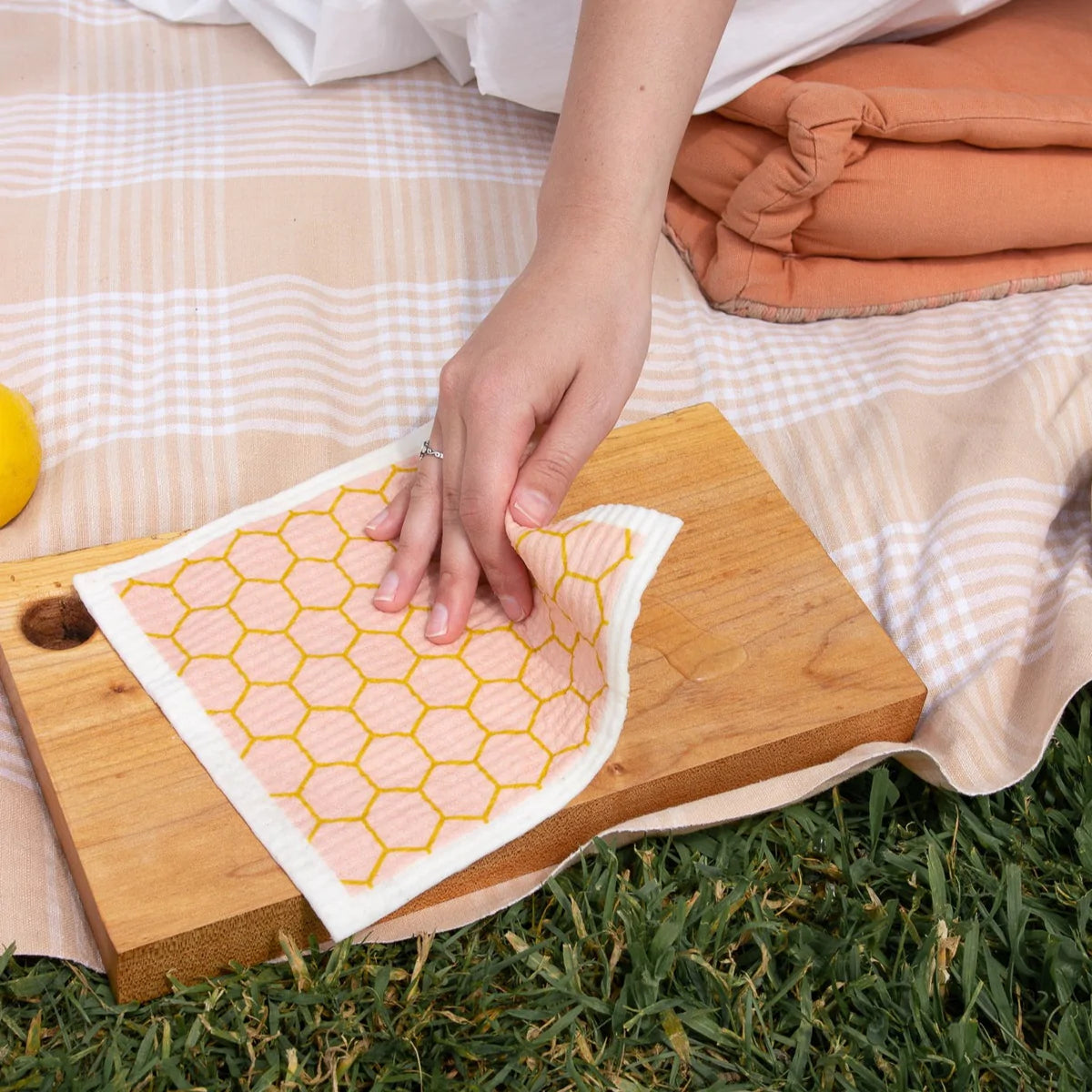 A woman's hand is wiping a wooden chopping board using the pink honeycomb Goldilocks dishcloth in a picnic setting.