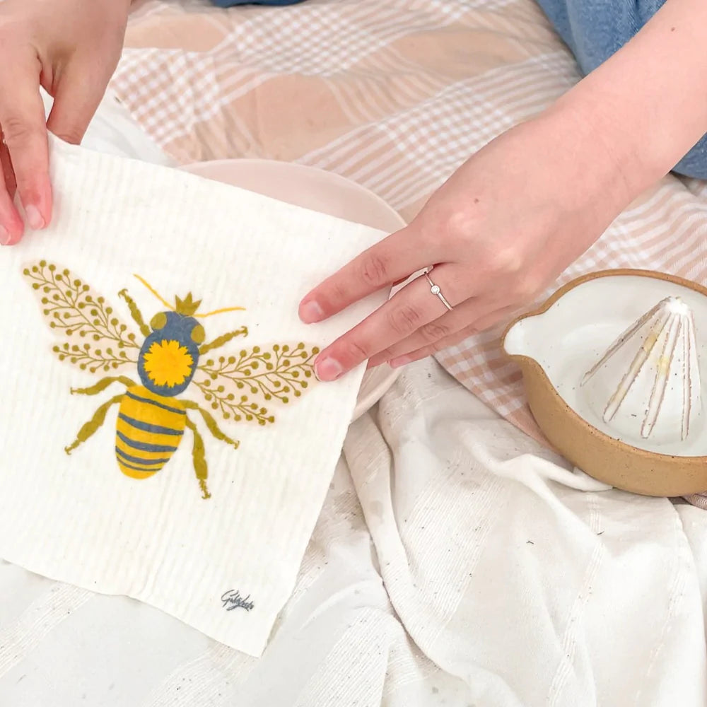 A woman's hand is holding the goldilocks queen bee dishcloth in a picnic setting.