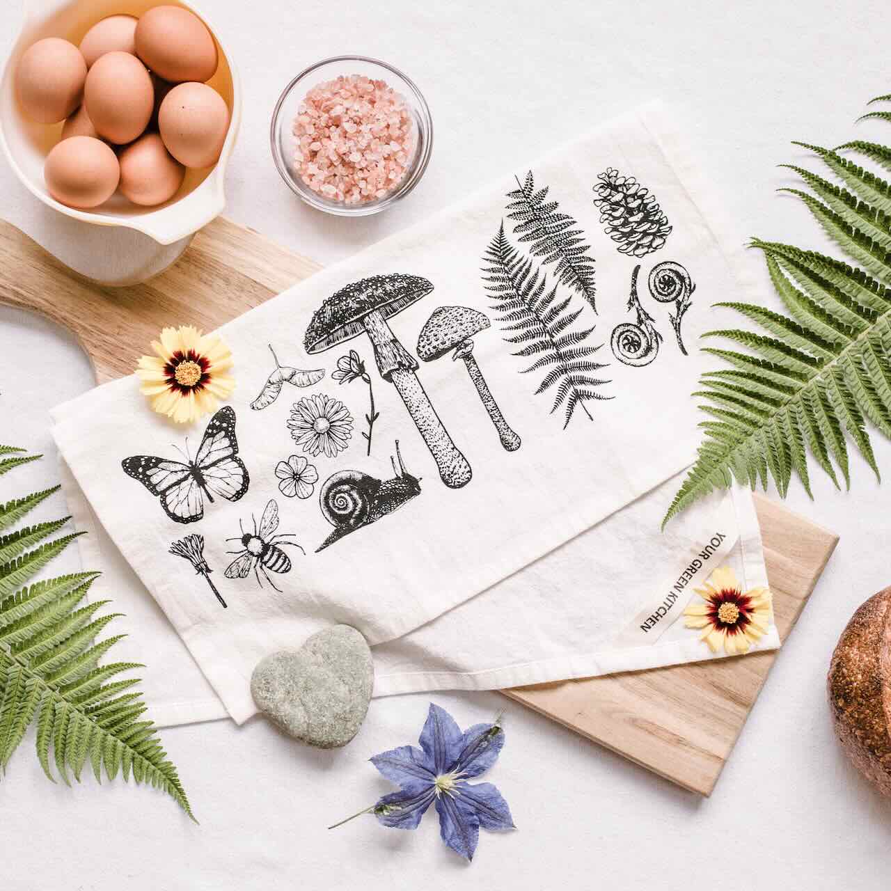 A white kitchen tea towel with a selection of plants, animals that are typically associated with an autumn forest, printed on, laying casually on top of a wooden chopping board, with pink sea salt, eggs and blue flower as decoration pieces around it.