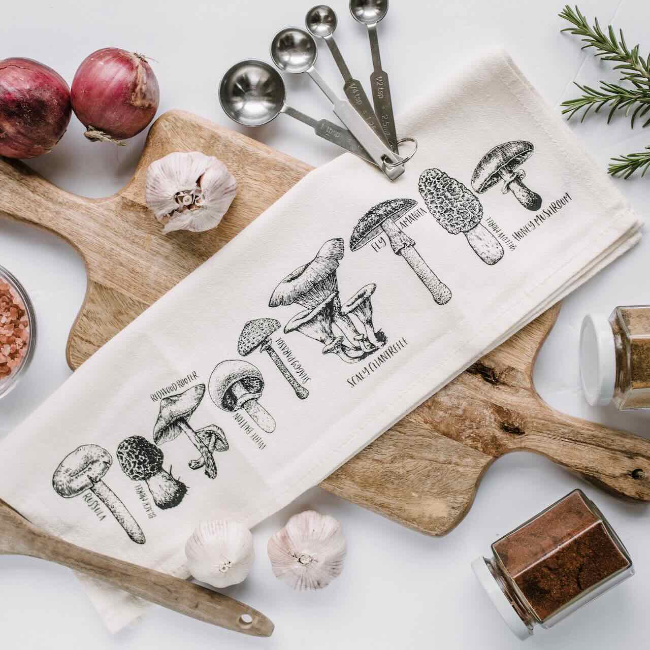A white kitchen tea towel with 9 different mushroom types printed, laying casually on top of a wooden chopping board, with onions, garlic, and some spices in  bottle as decorating items.