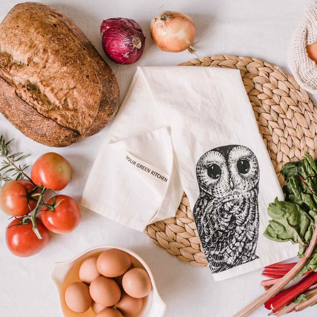 A white kitchen tea towel with a large owl printed, laying casually on a white table top, surrounded by bread, red tomatoes on vine, eggs and onions.