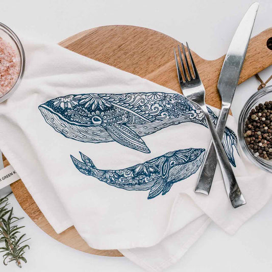 A white kitchen tea towel with two blue whales (a mother and her offspring) printed, laying casually on top of a wooden chopping board, next to a pair of fork and knife.