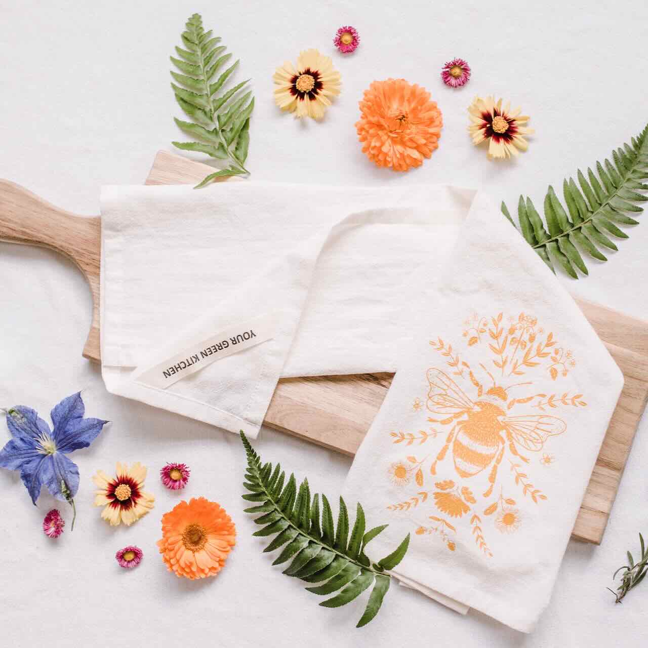 A white kitchen tea towel with a large yellow bee printed, laying casually on top of a wooden chopping board, with flower and leaves as decoration pieces around it.