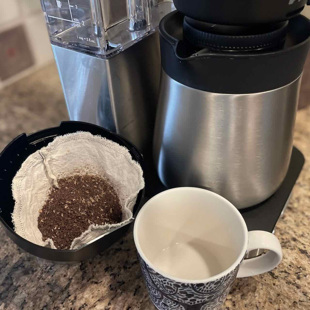 size 4 coffee filter filled with ground coffee inside basket next to the coffee maker and a porcelain coffee cup