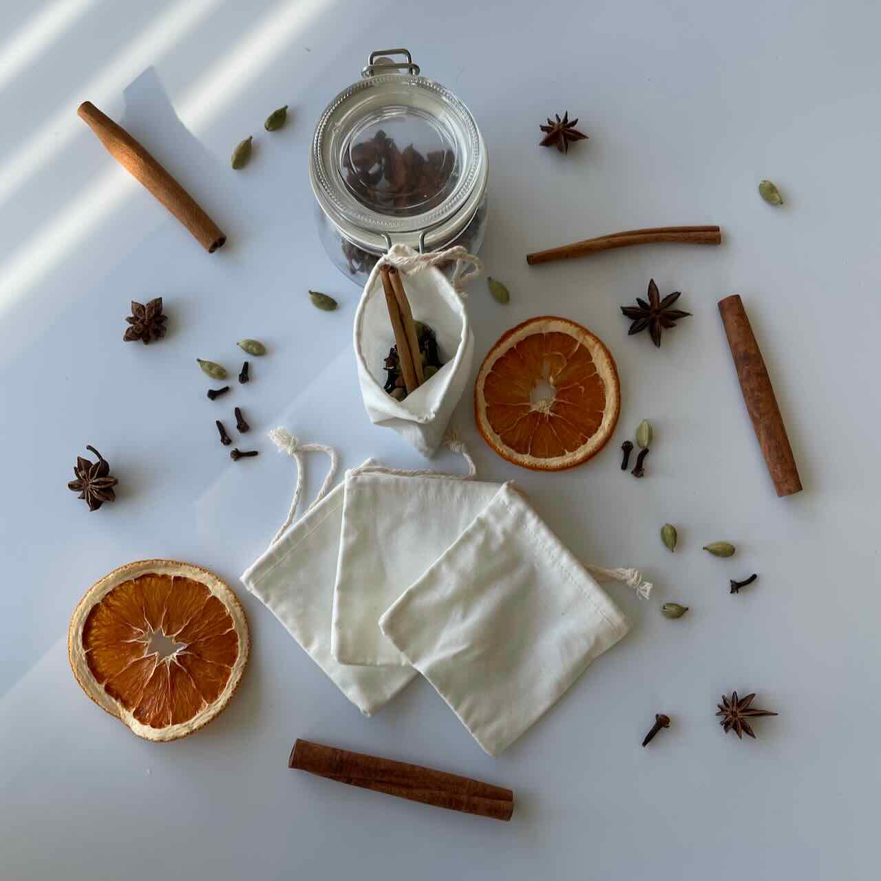 reusable muslin tea bags lay on top of a white table with spices and dried orange slices scattered around them