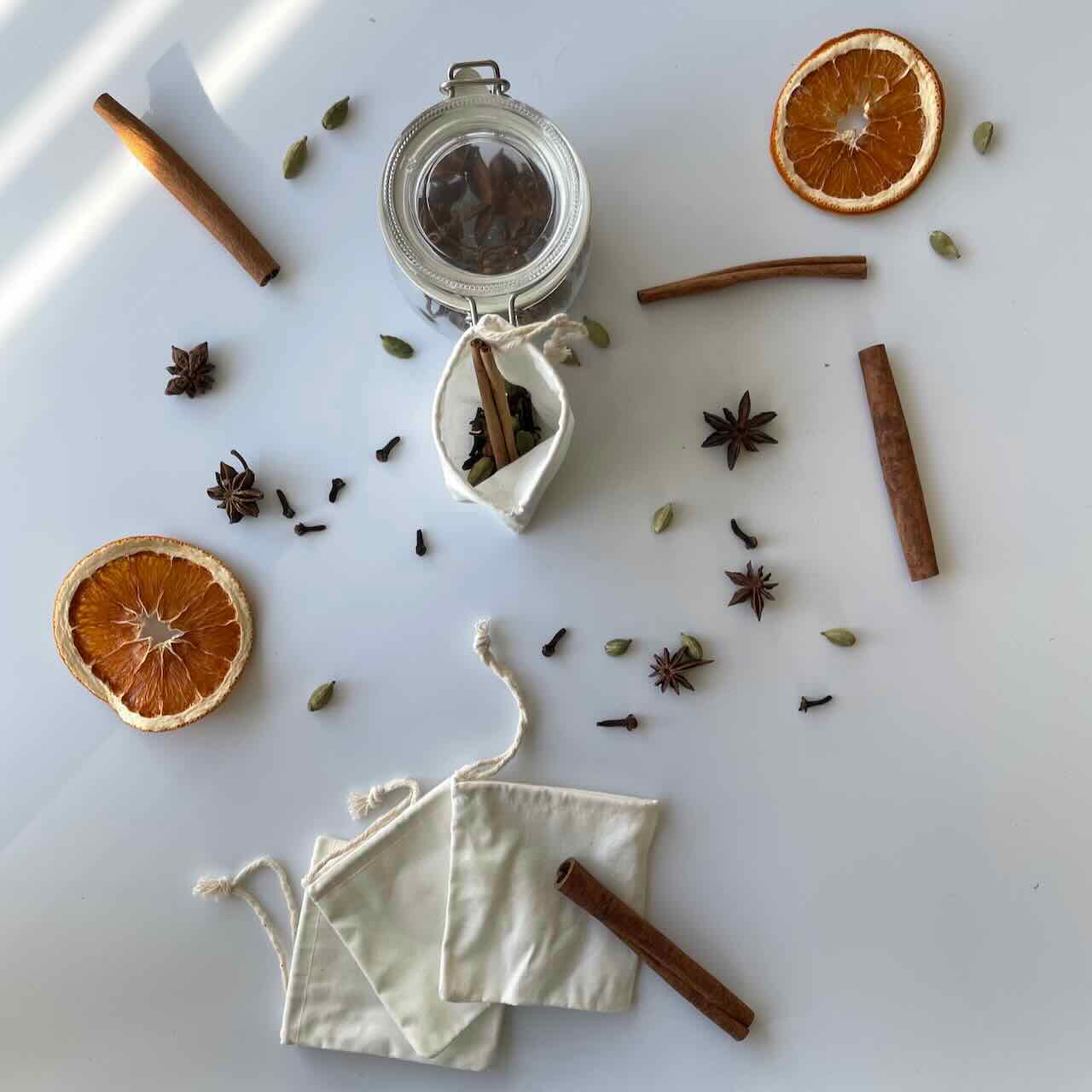 reusable muslin tea bags lay on top of a white table with spices and dried orange slices scattered around them