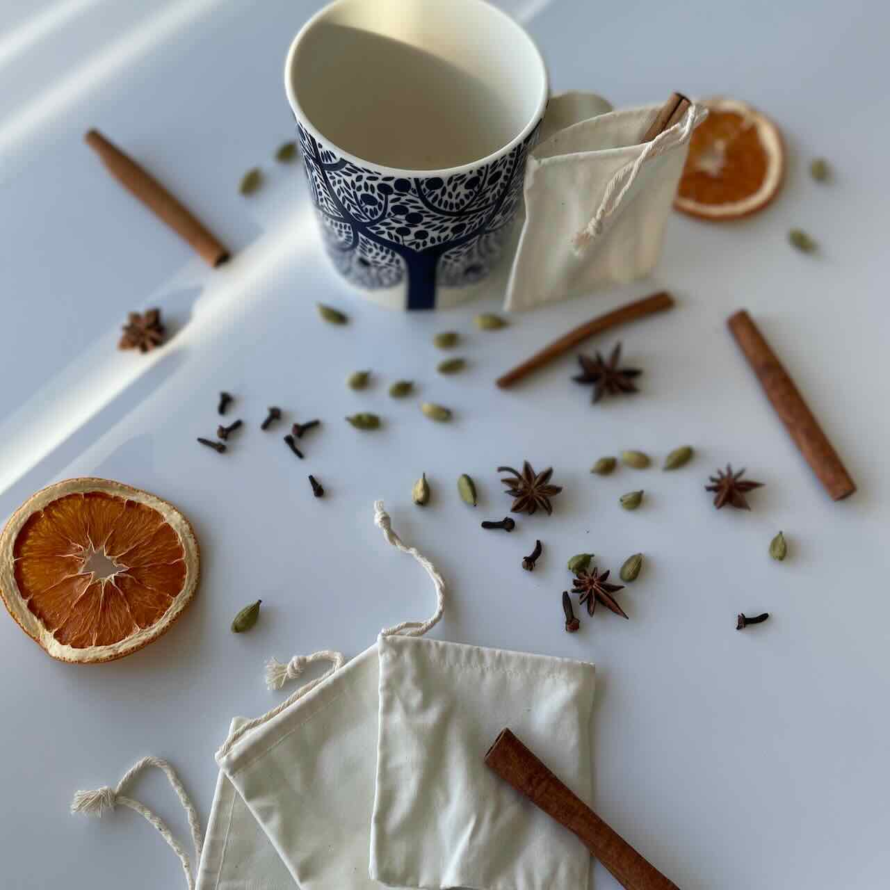 reusable muslin tea bags lay on top of a white table with tea mug, spices and dried orange slices scattered around them