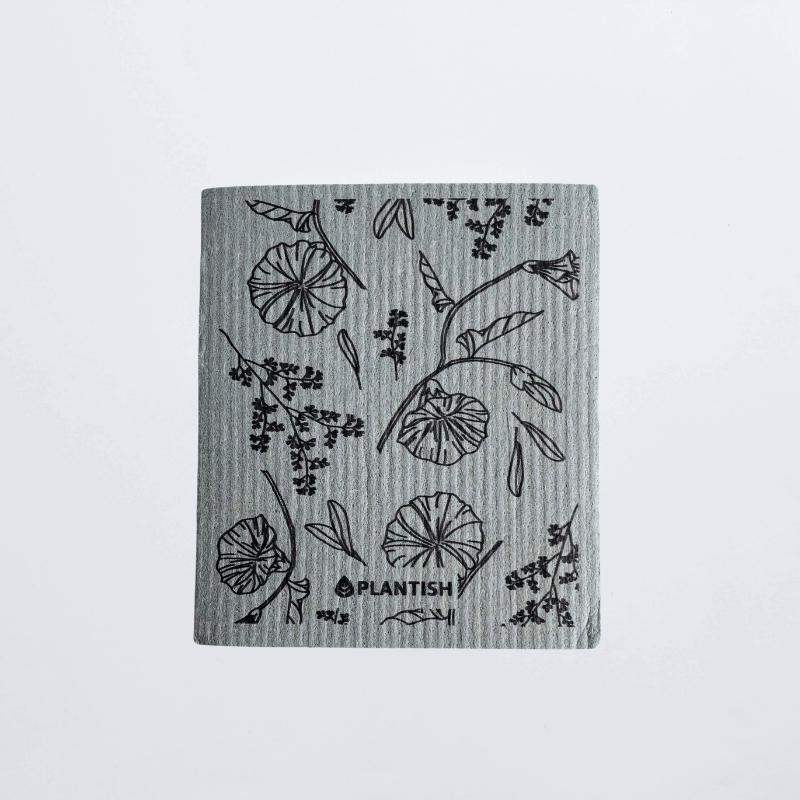 Swedish dish cloth by Plantish lay flat on a white table top. The dish cloth is grey coloured with morning glory sketch as pattern. 