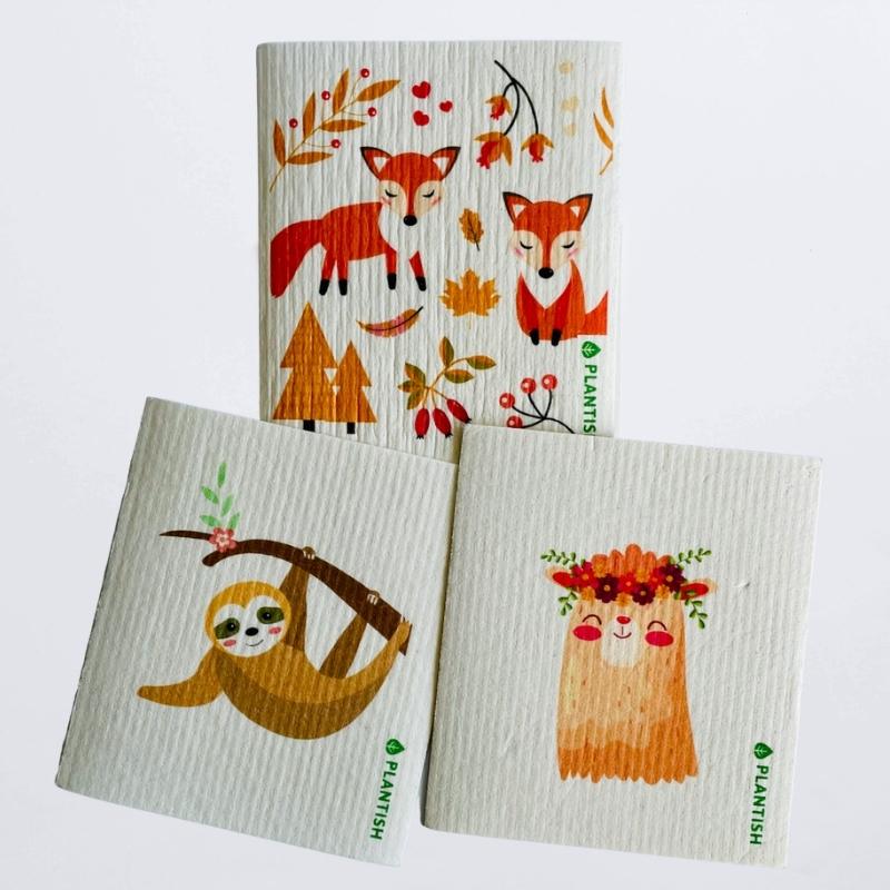 Set of 3 swedish dish cloths on a white table top. The patterns are inspired by cute and wild animals: money hanging on a tree, smiling llama and resting foxes
