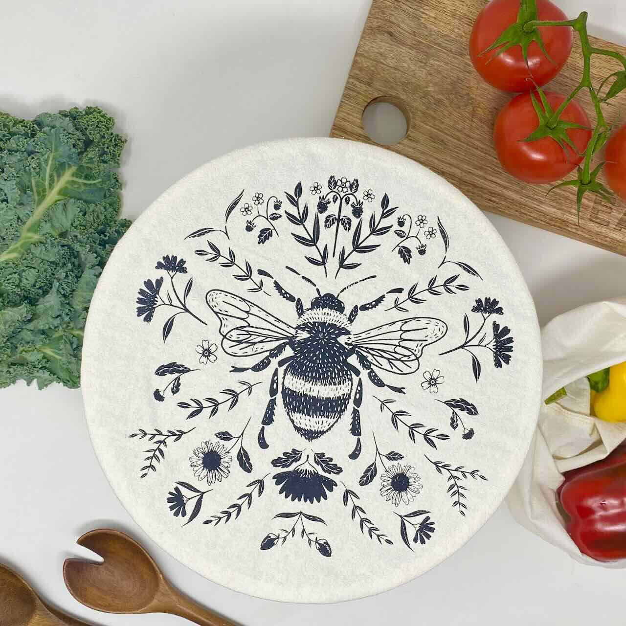 Flat lay of a large bowl covered by a cotton charcoal bee patterned bowl cover on a wooden chopping board, surrounded by vegetables such as red tomatoes, kale and peppers.