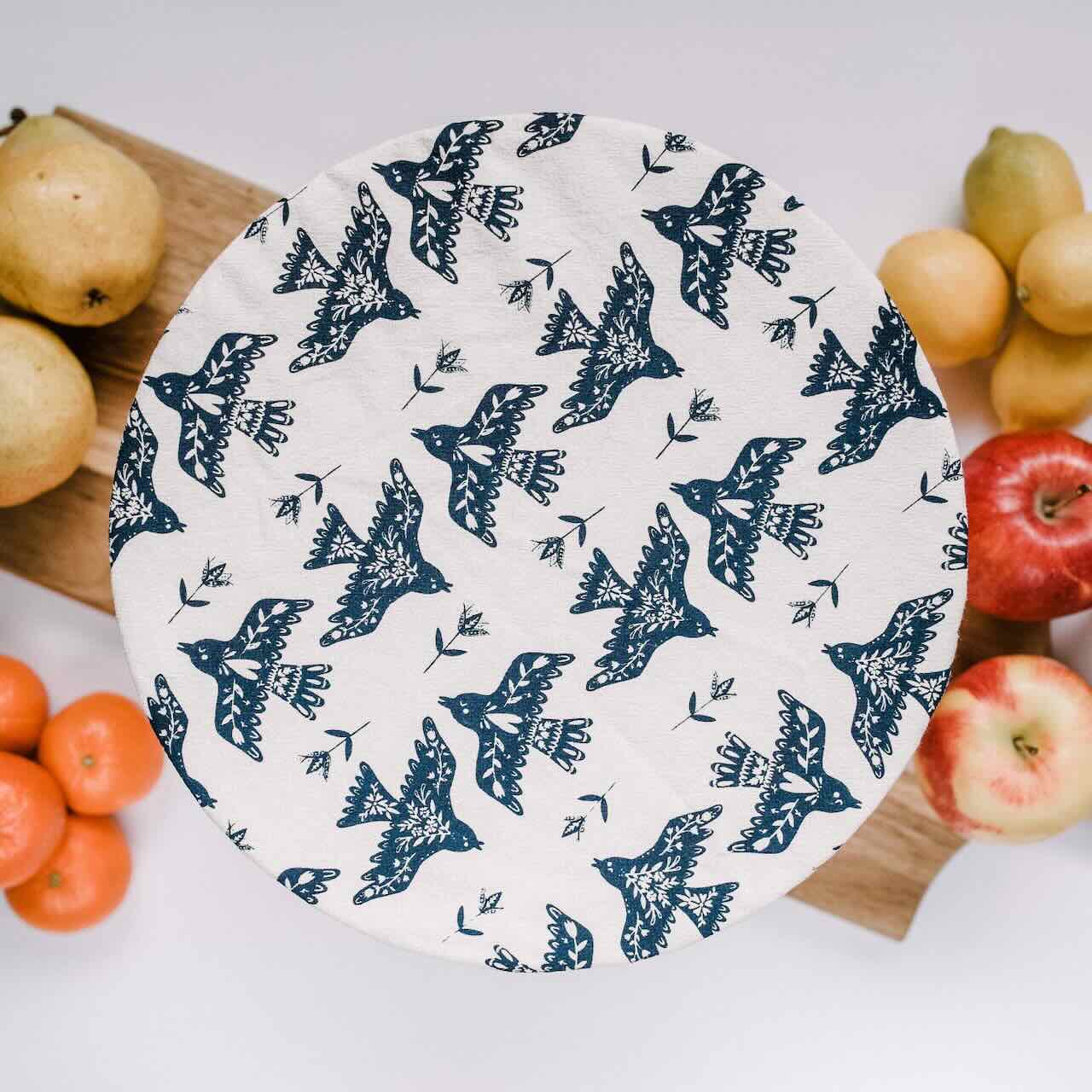 flat lay of a large bowl covered by a navy flying birds patterned cotton bowl cover on a wooden chopping board, surrounded by apple, oranges and lemons.