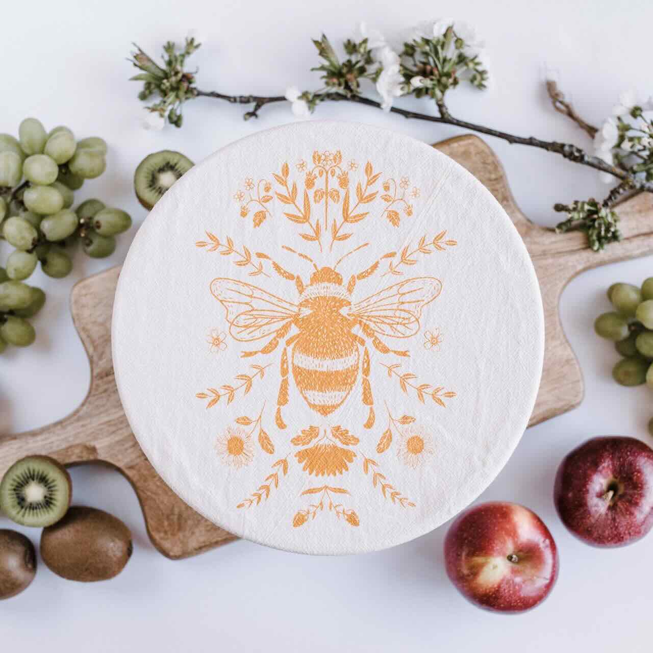 Flat lay of a medium sized bowl covered by a cotton yellow bee patterned bowl cover on a wooden chopping board, surrounded by fruits such as apples, grapes and kiwi.