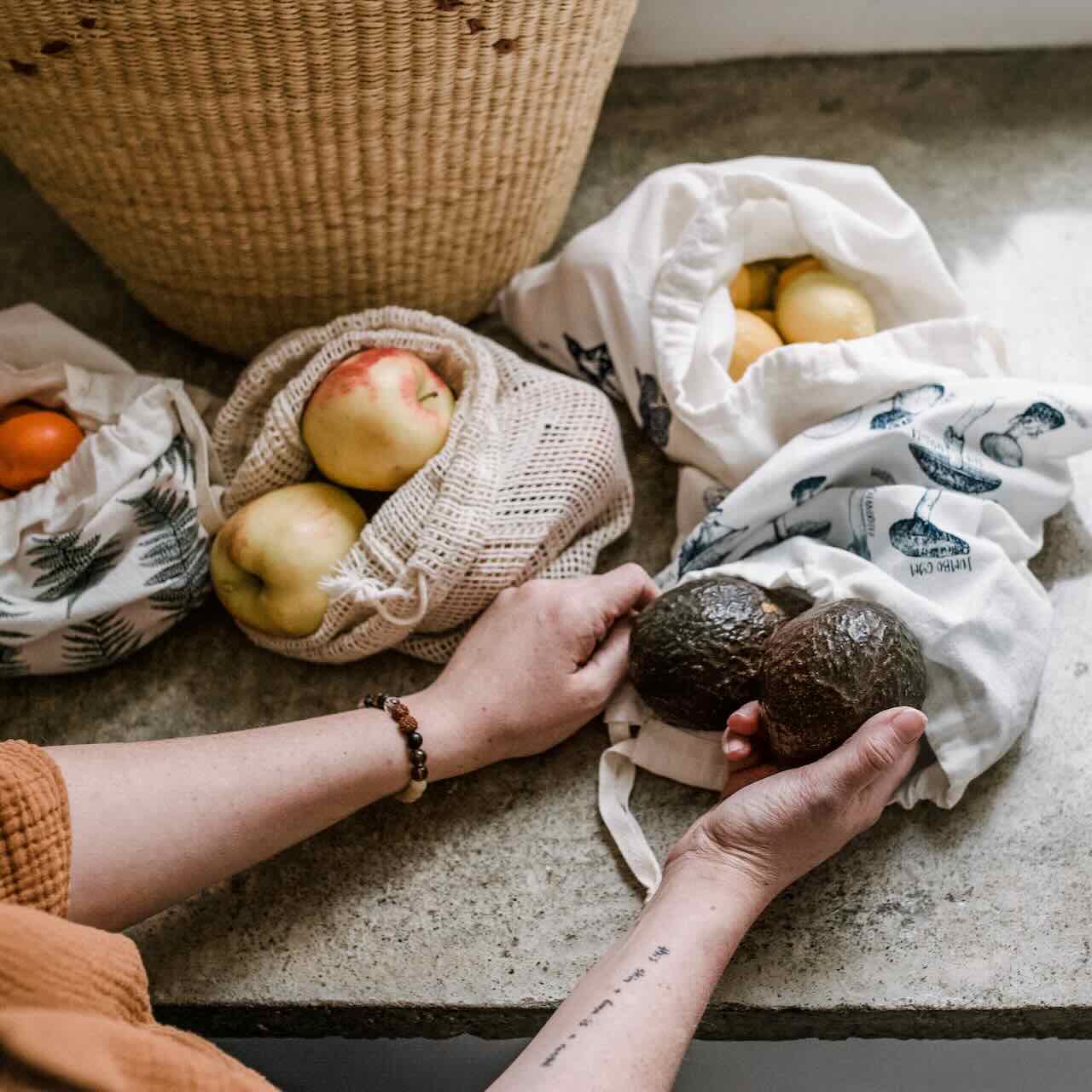 A woman putting avocado into a mushroom patterned produce bag on a concrete style counter top.