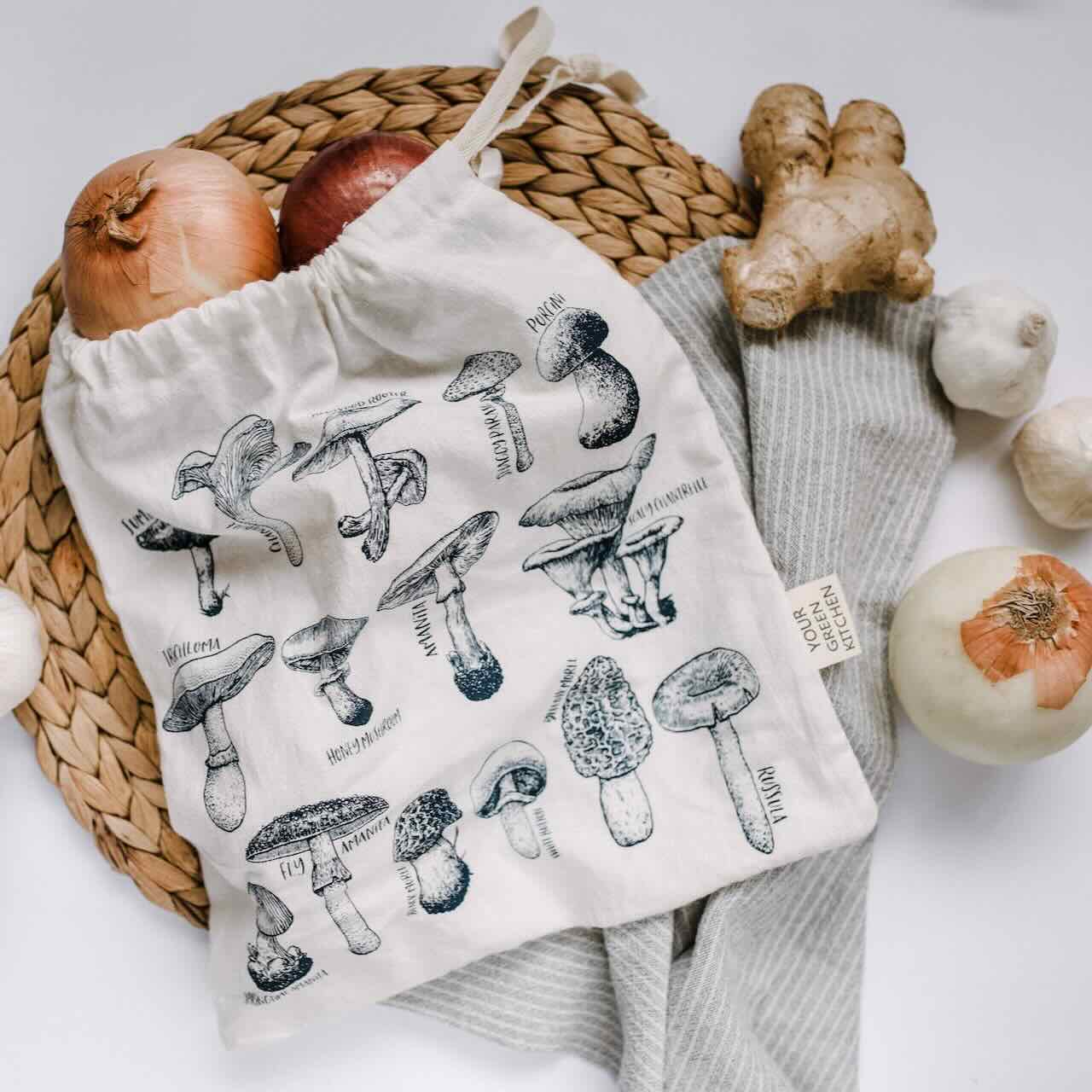 A produce bag with varietal of mushroom patterns laying flat on a rattan placemat, with onions and ginger, and garlic scattered around it.