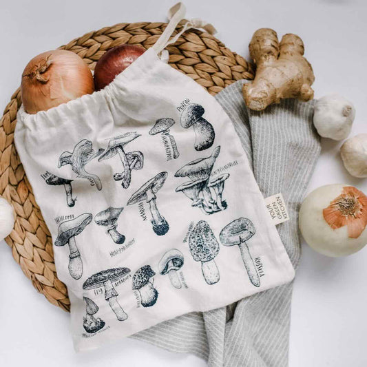 A produce bag with varietal of mushroom patterns laying flat on a rattan placemat, with onions and ginger, and garlic scattered around it.