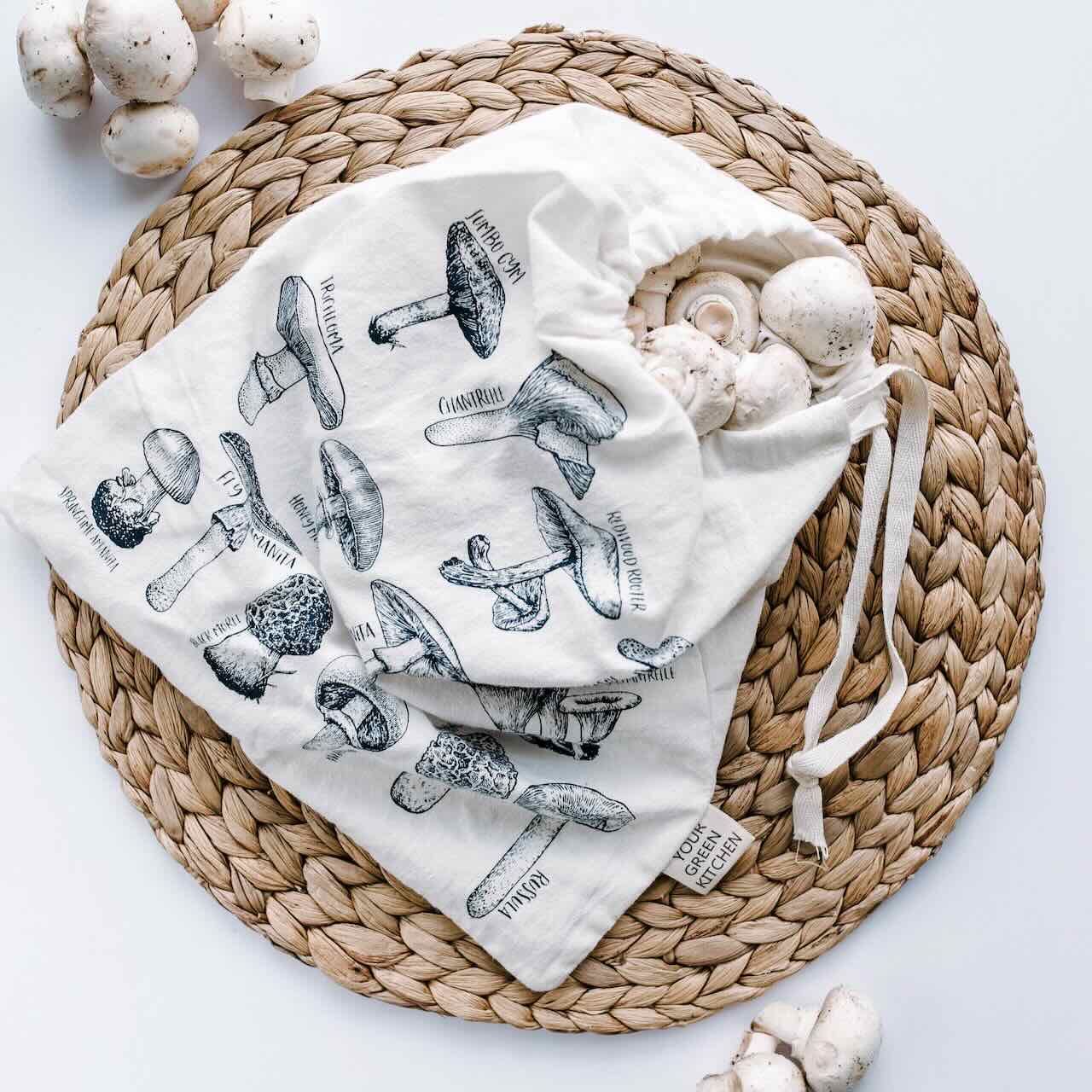 A produce bag with varietal of mushroom patterns laying flat on a rattan placemat, with mushrooms scattered around it.