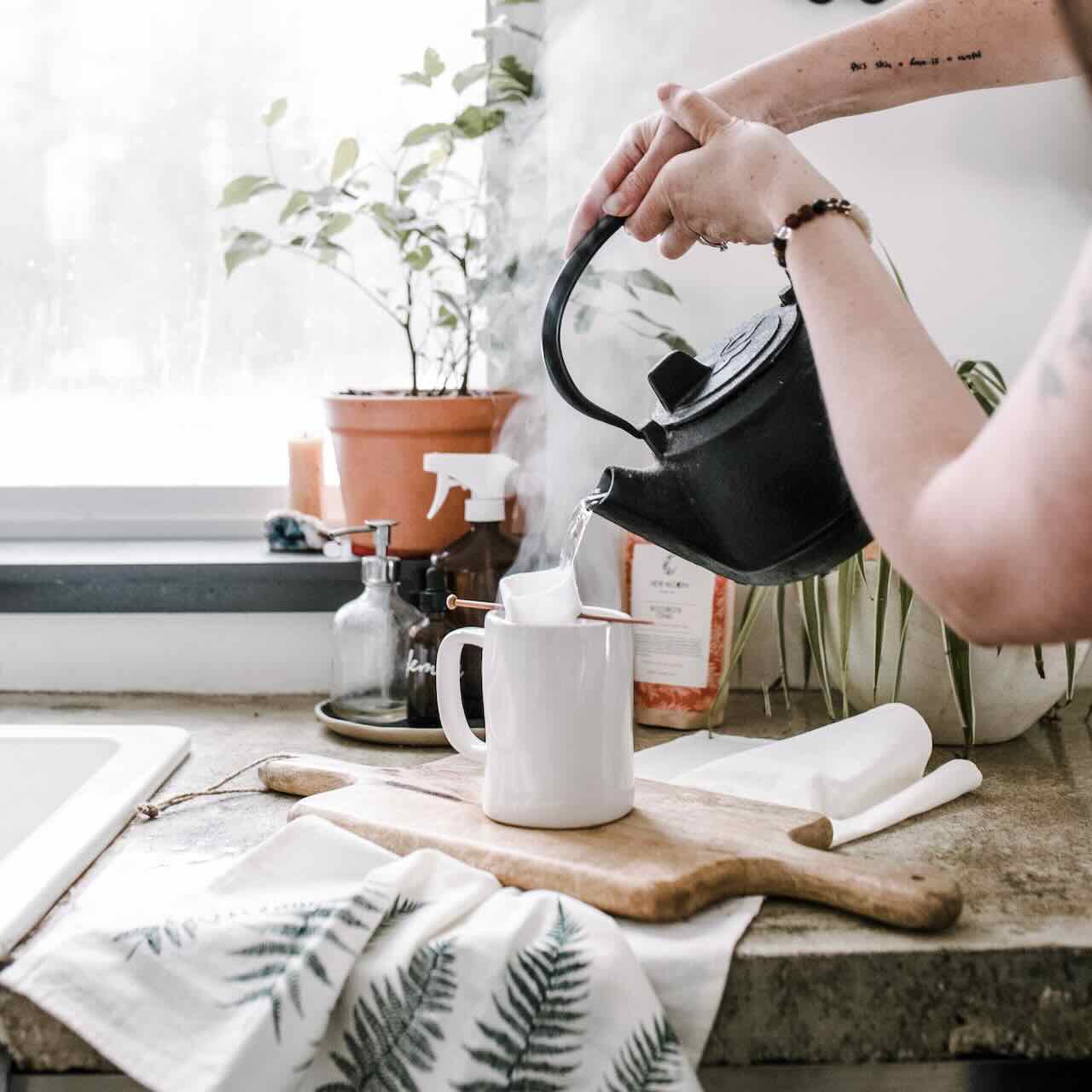 A woman pouring water into a white mug with reusable tea bag from a cast iron kettle.