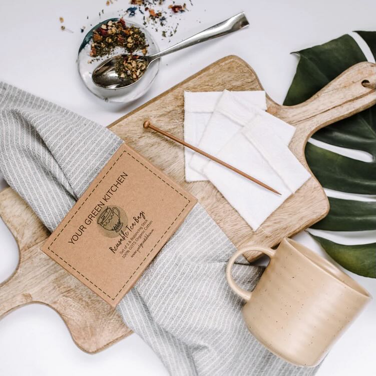 3 white reusable tea bag with its balancing stick, and its paper box on a wooden chopping board. Scattered around it is a earth tea mug, cotton tea towel and some loose tea on a spoon.