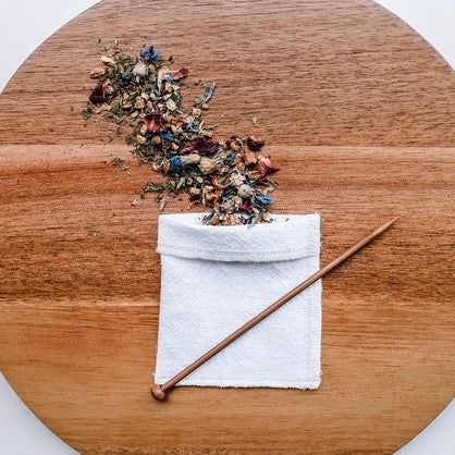 One white reusable cotton tea bag, loose floral tea leaves, and the balancing stick on a walnut chopping board.